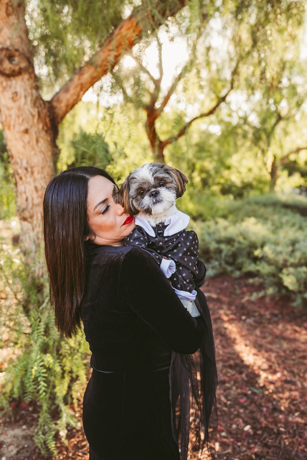 A small fluffy dog wearing polka dots gets affection while being held in a wooded outdoor setting at sunset.