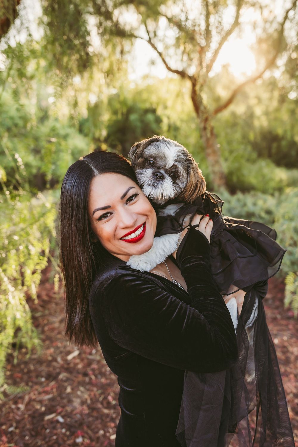 A woman in a black dress holds a small Shih Tzu dog wearing a fancy black outfit in a sunlit outdoor setting.