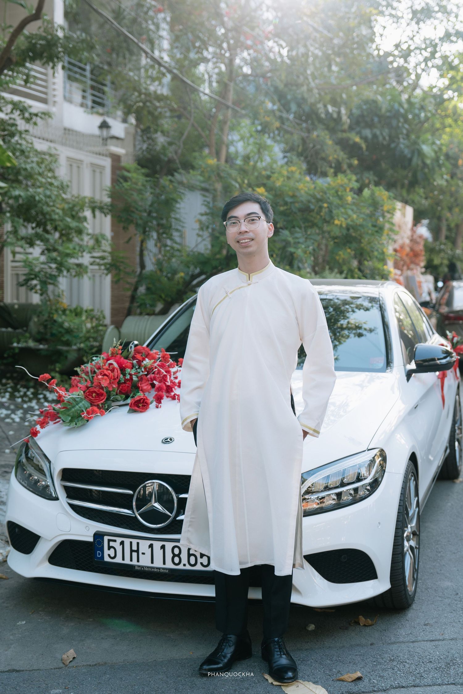 Vietnamese groom in white ao dai smiling beside white Mercedes wedding car with red floral decoration.