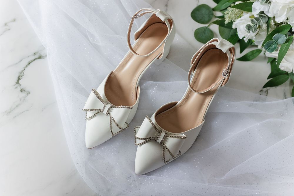 Elegant white leather bridal shoes with pointed toes and bow details displayed against a light background with florals.