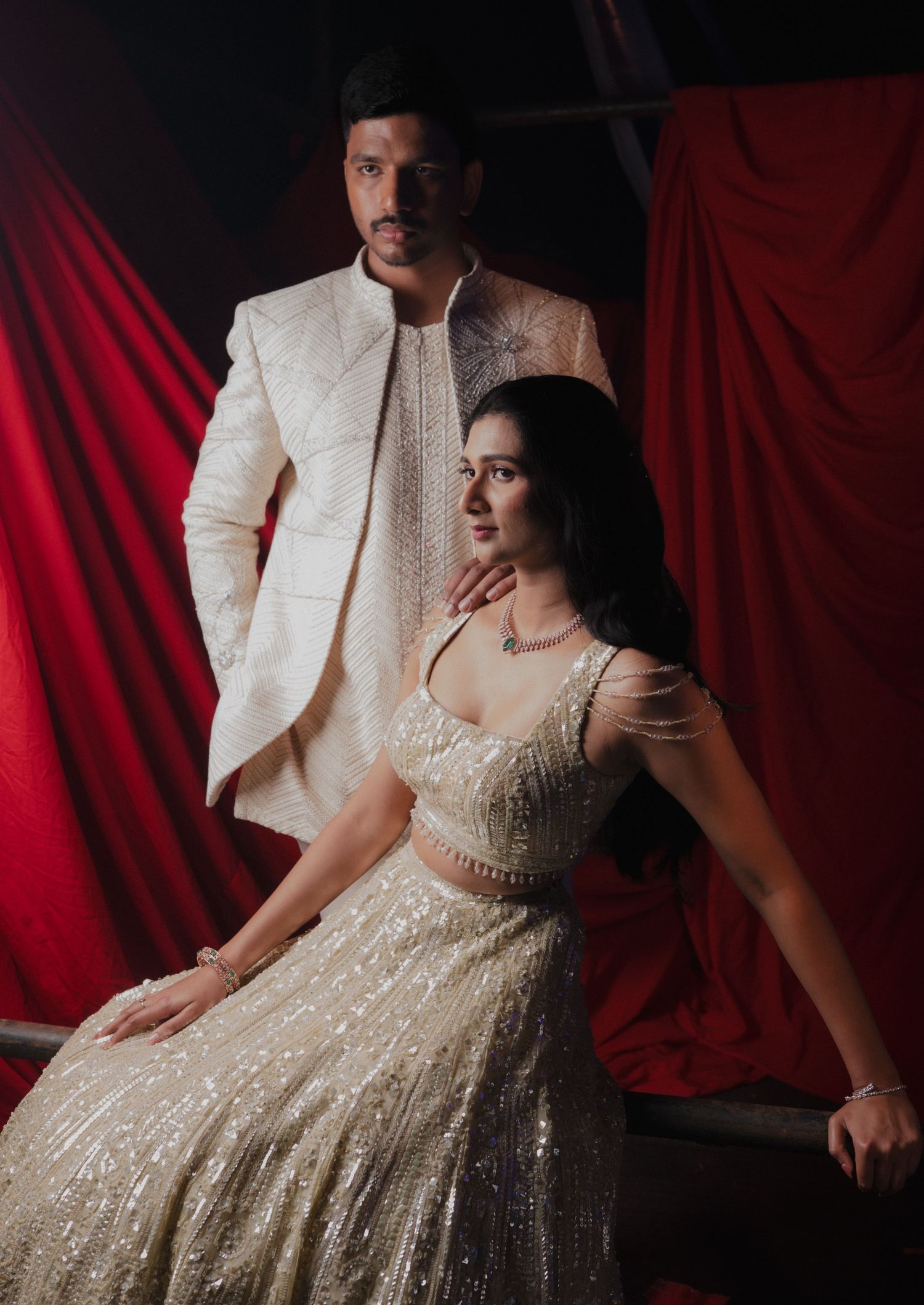 Elegantly dressed couple in matching cream and white traditional Indian formal attire against red backdrop.