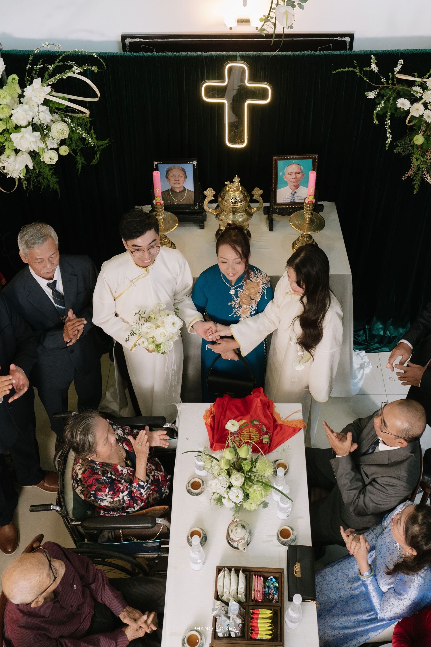 Vietnamese Christian wedding ceremony with couple exchanging rings at altar decorated with cross, flowers, and ancestral portraits.