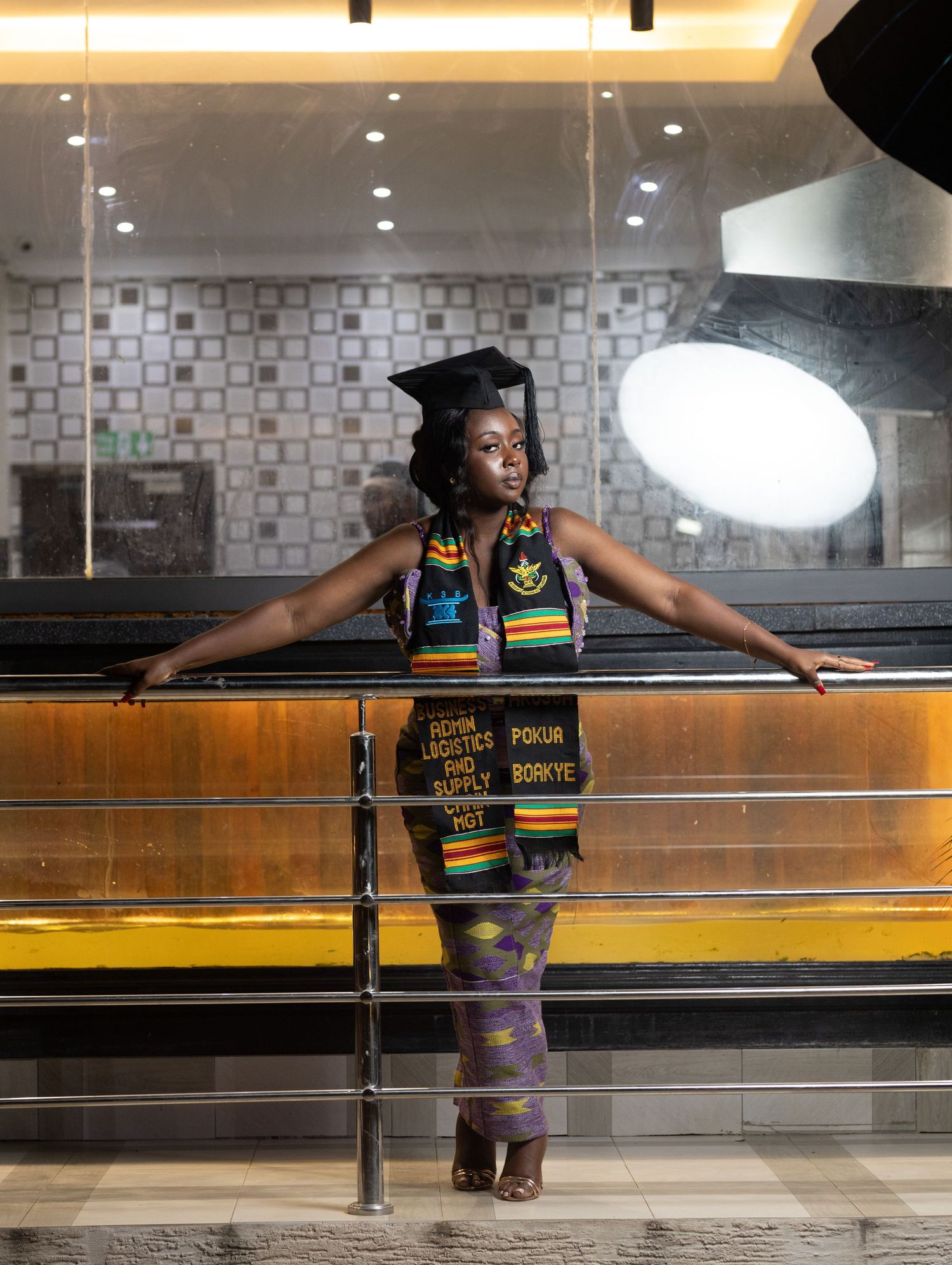 Graduate wearing academic regalia poses against wooden railing in modern interior space with dramatic lighting.
