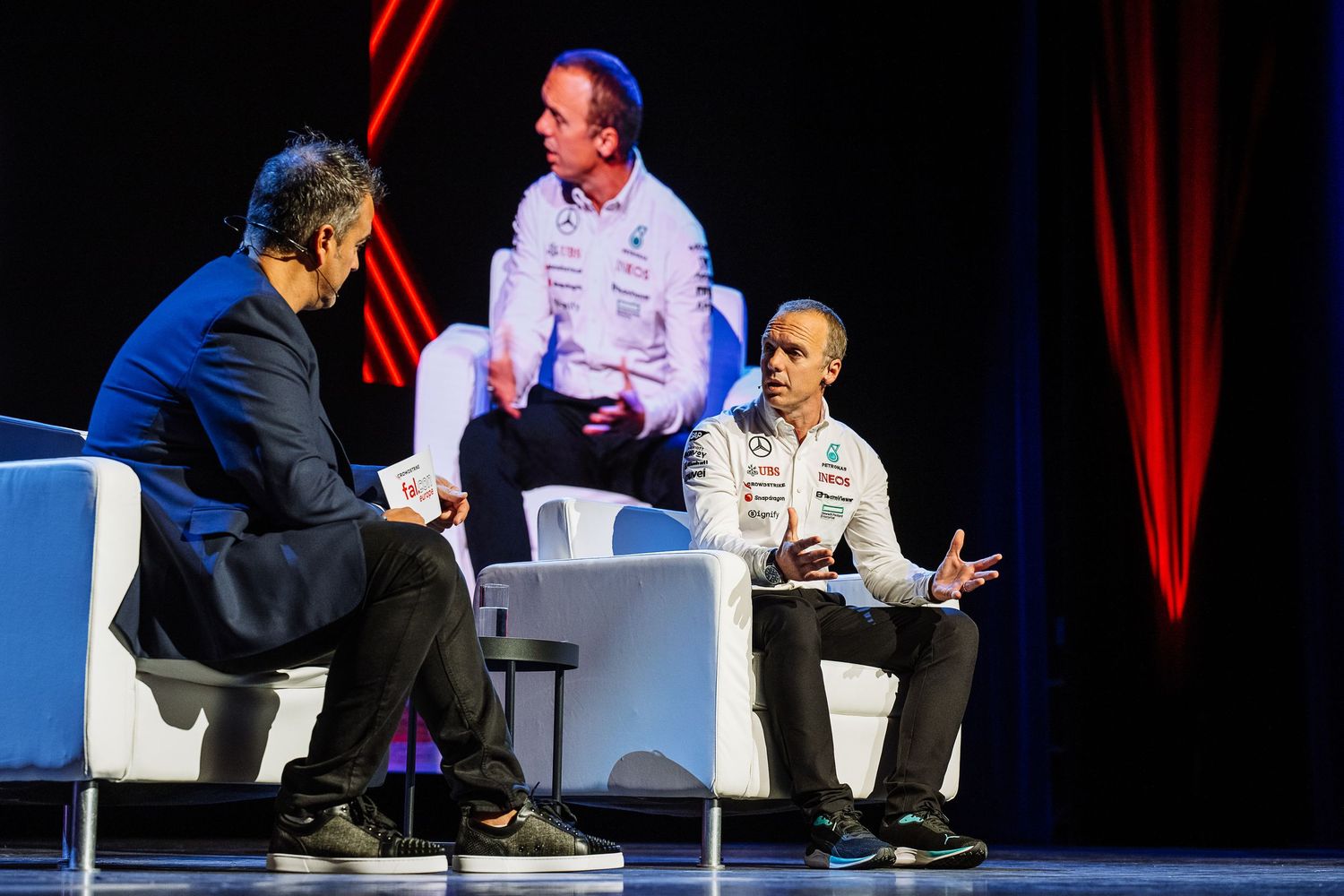 Business conference speakers engage in discussion on stage with red curtain backdrop.
