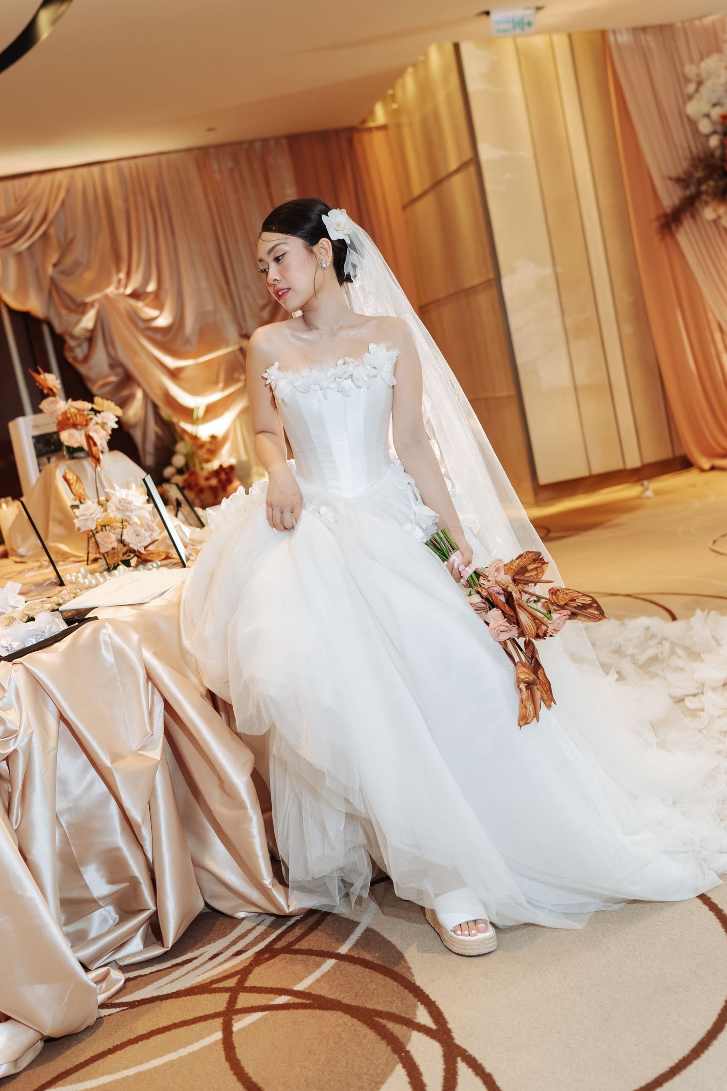 Bride in white ballgown wedding dress with veil holding bouquet in decorated reception hall.
