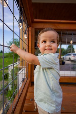 Baby in light blue shirt exploring and playing on a wooden deck railing.