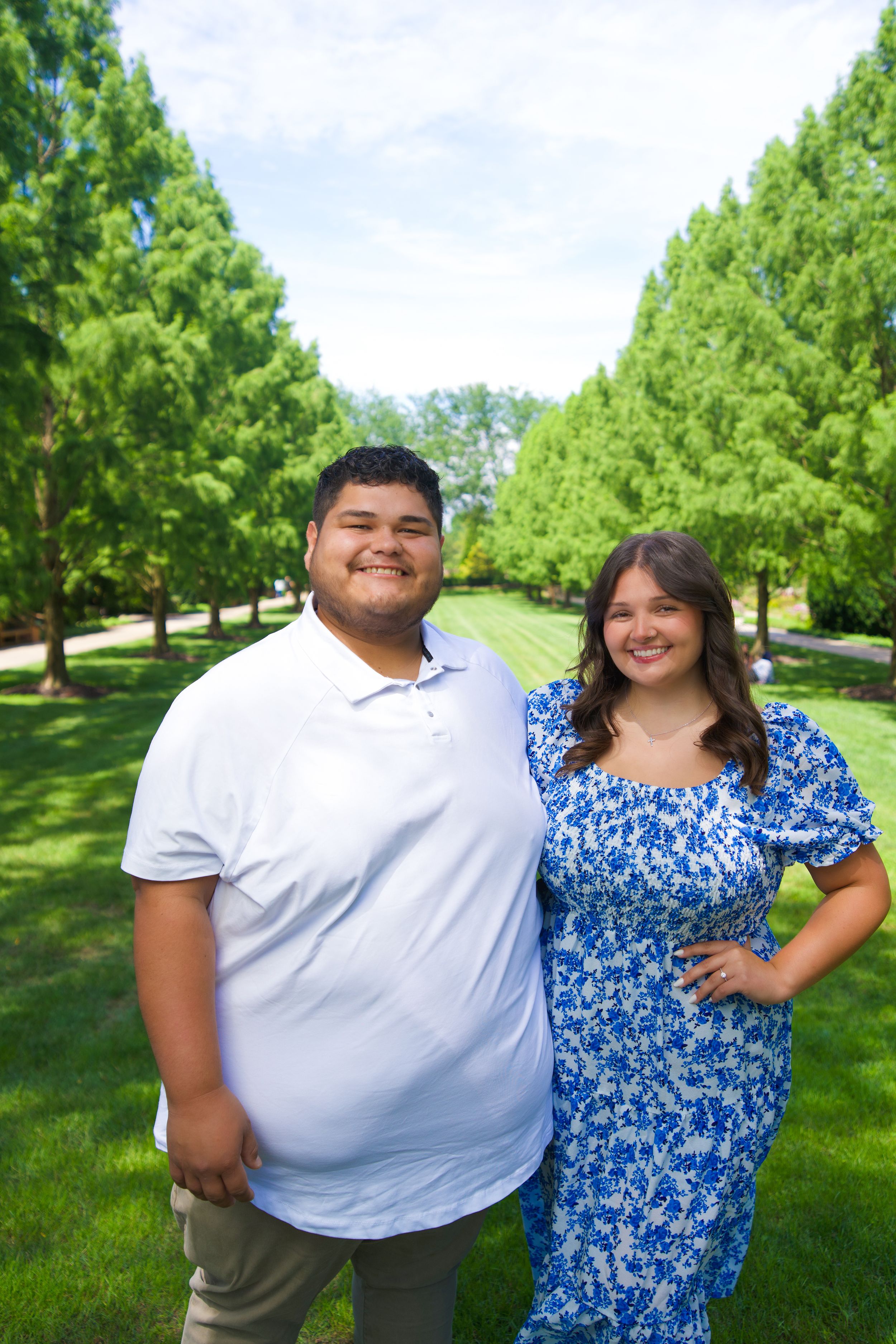 A couple poses together outdoors in a green park setting on a sunny day.