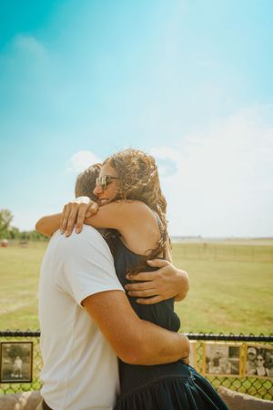 Two people embrace warmly against a bright blue sky and grassy field backdrop.