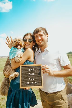 A couple poses with their dog and a letterboard announcing their engagement date.