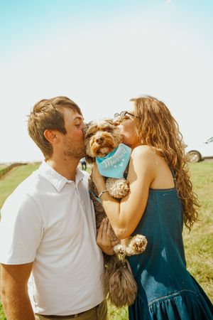 A couple shares champagne while holding their small dog in the sunlight.