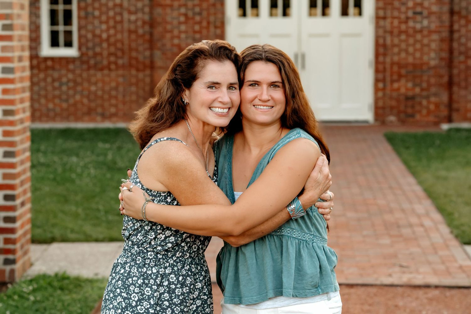Two people in casual summer outfits embrace while standing on a brick pathway near a traditional brick building.