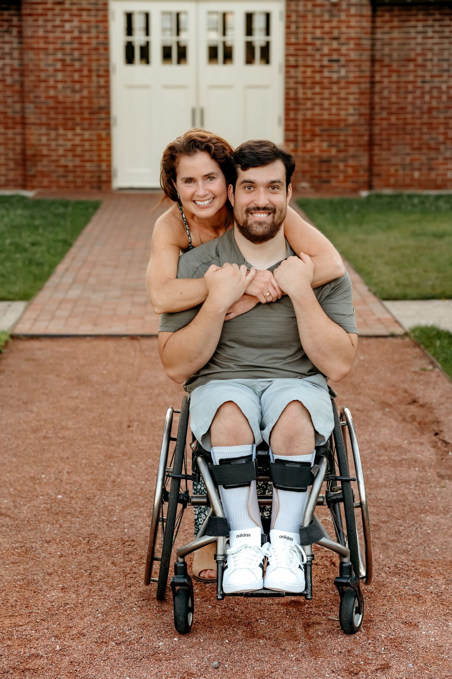 Two people share a loving embrace on a brick path while one sits in a wheelchair, smiling together outside a brick building.