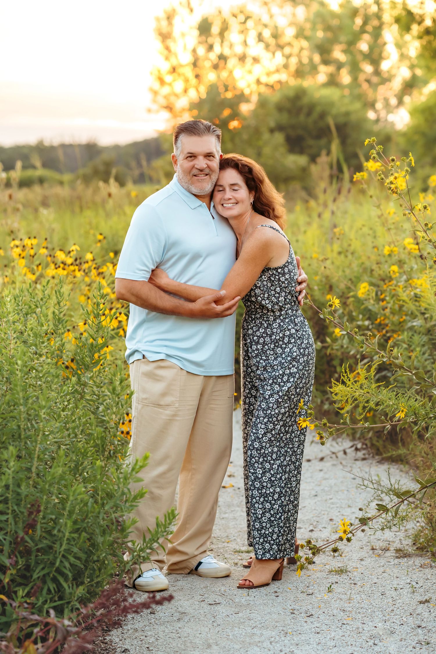 A couple embraces lovingly among blooming yellow wildflowers on a sandy path during a summer evening outdoors.