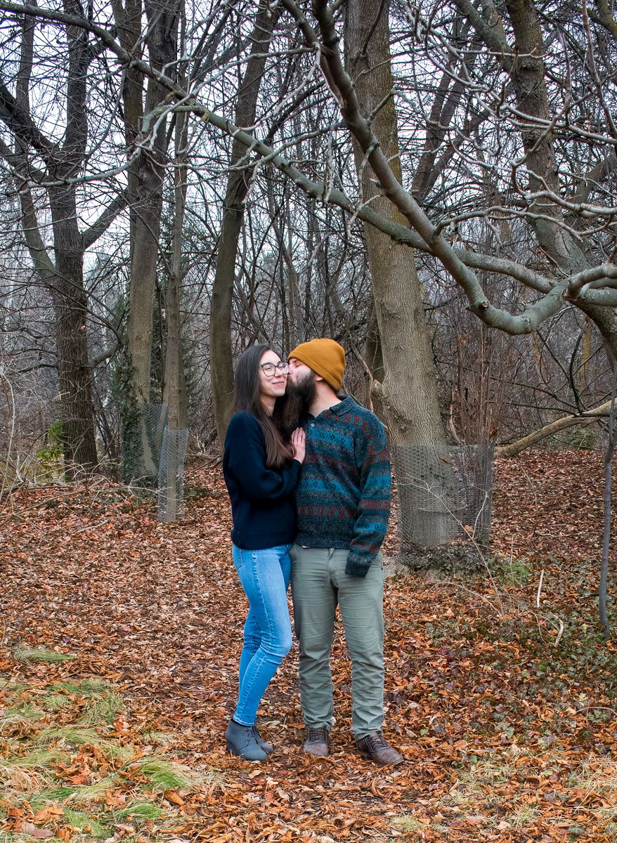 A couple shares intimate moments in a wooded area covered with fallen autumn leaves.
