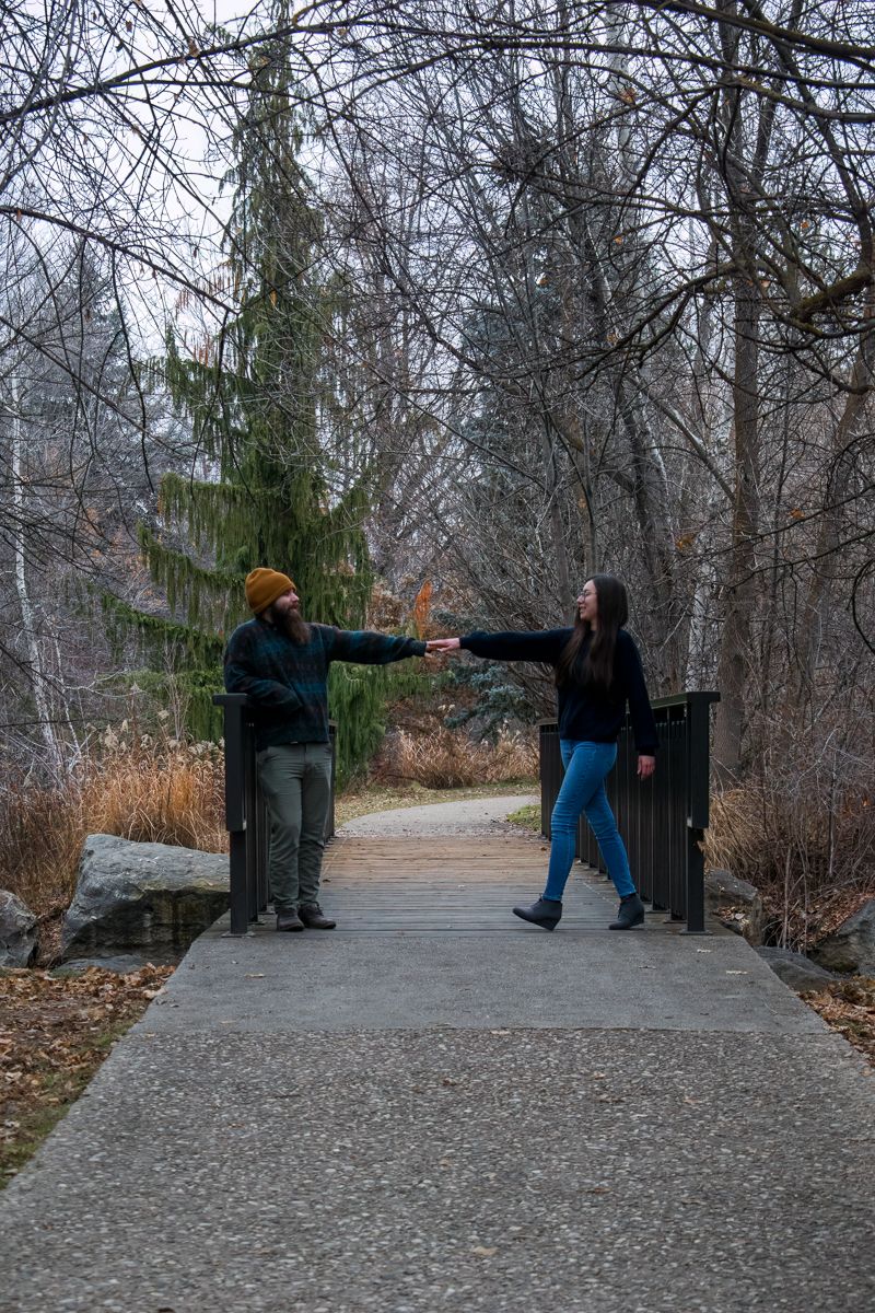 Two people greet each other with outstretched arms on a path in a winter forest setting.