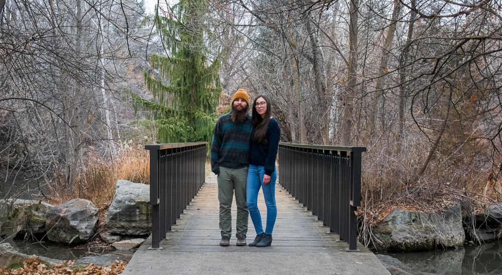 People walking across a wooden footbridge through a winter forest landscape with bare trees and snow covered ground.
