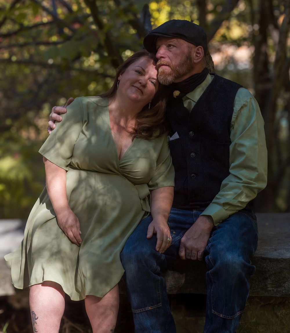 A couple in vintage-style clothing share an intimate moment together on a stone wall in a wooded setting.