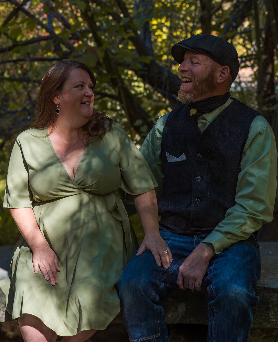 A couple shares a tender moment together while sitting outdoors in dappled sunlight among trees and foliage.