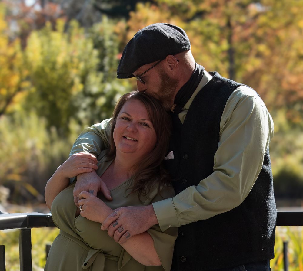 A couple shares a tender embrace outdoors on a sunny autumn day with fall foliage in the background.