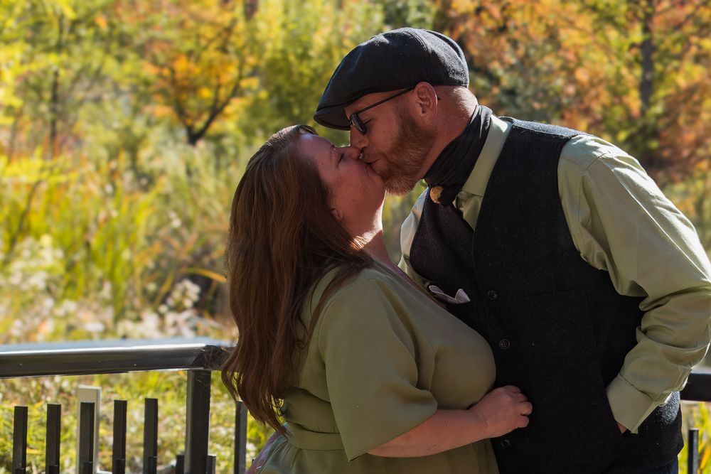 A couple shares a romantic kiss in a garden setting with autumn foliage and a wooden fence in the background.