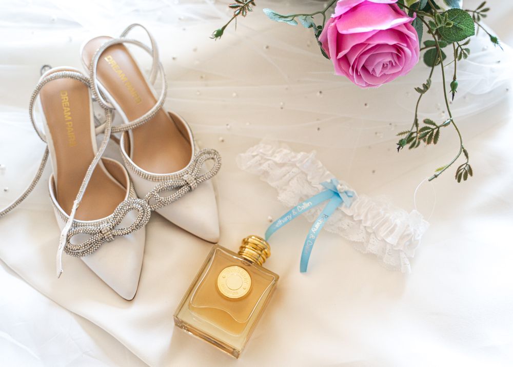 Wedding accessories including silver heels, perfume bottle, and pink rose arranged on white fabric.
