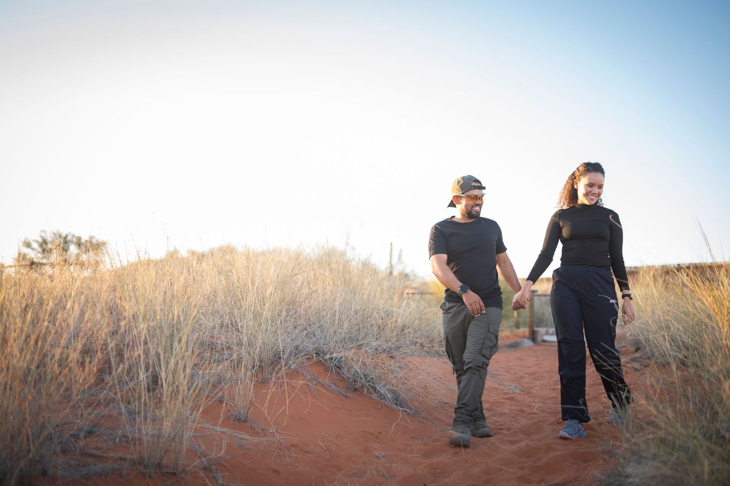 Two people walk through tall golden grass in a field during sunset with a blue sky overhead.