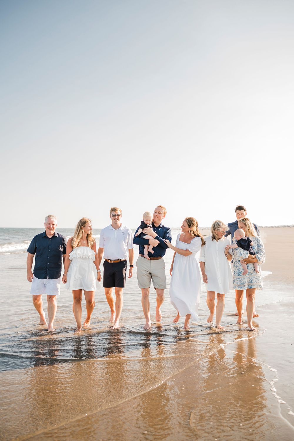 Family wearing coordinated white and navy outfits walking along the beach shoreline at sunset.