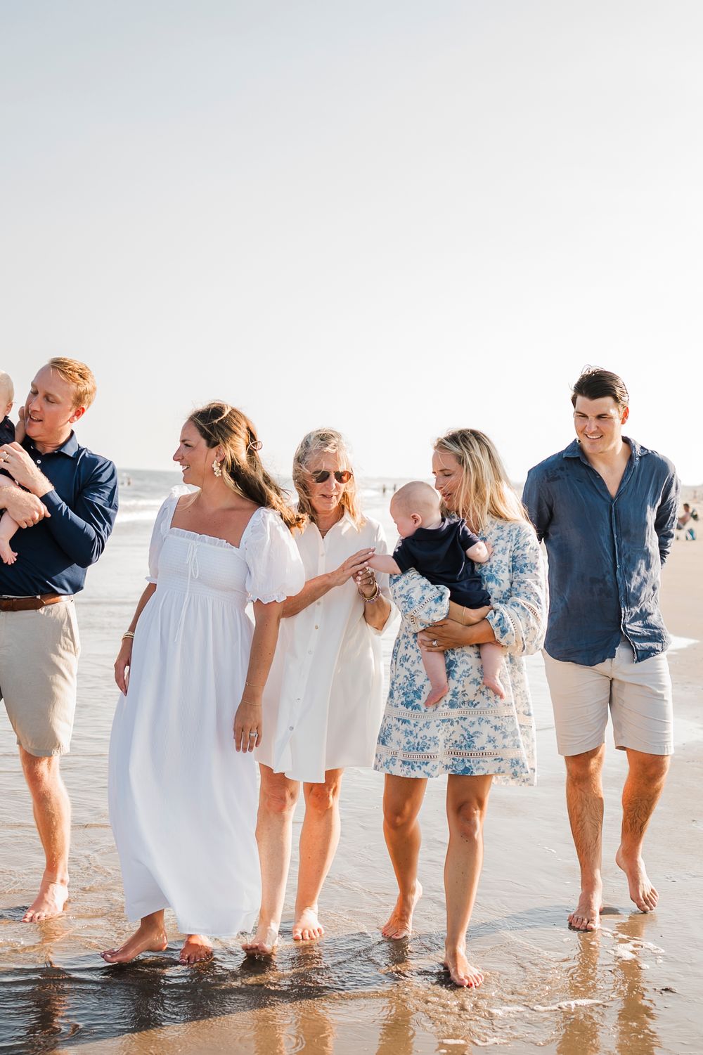 A group walking together barefoot along the beach shore in casual summer attire.