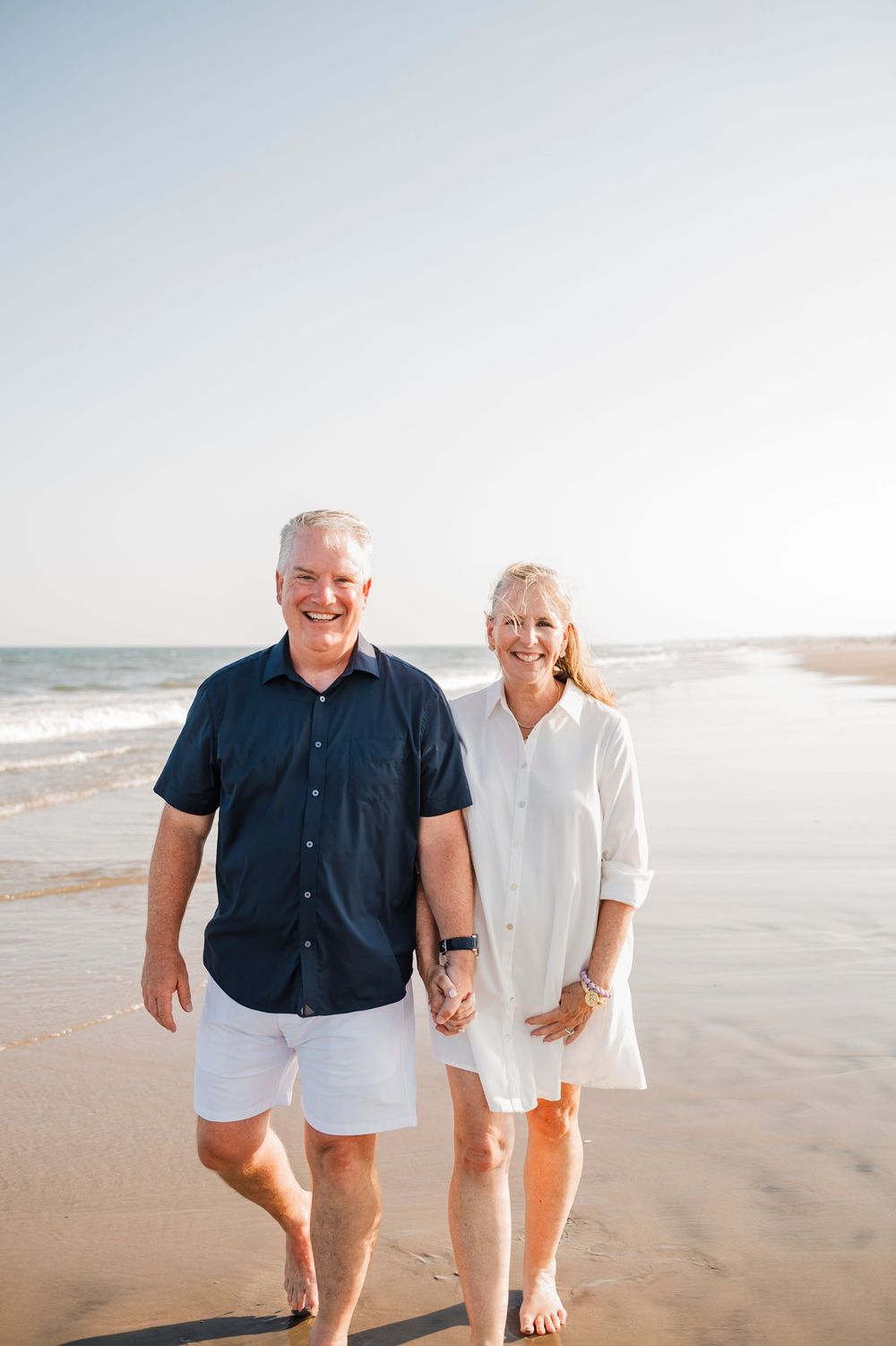 Couple walking and laughing together along a sunny beach wearing navy and white summer outfits.