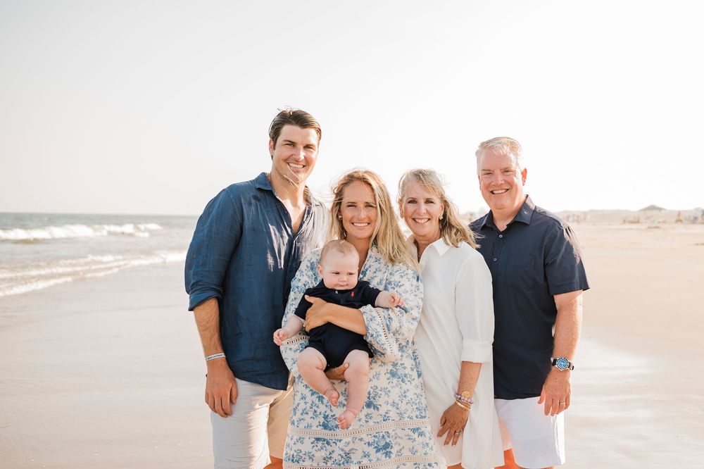 A family stands together smiling on a sunny beach with waves and sand in the background.
