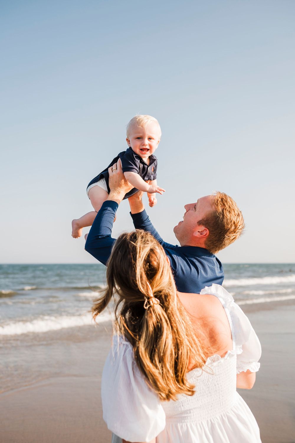 Joyful parents lifting and playing with their baby at the beach with waves in the background.