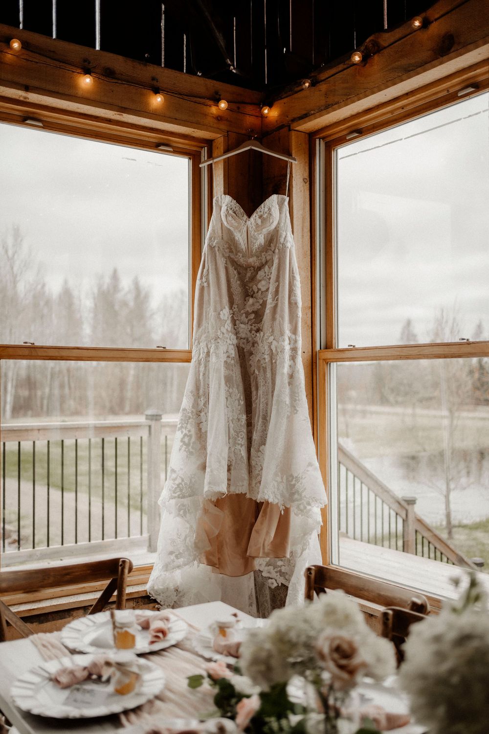 Wedding dress hanging in window with wooden furnishings and misty forest view creating romantic atmosphere.