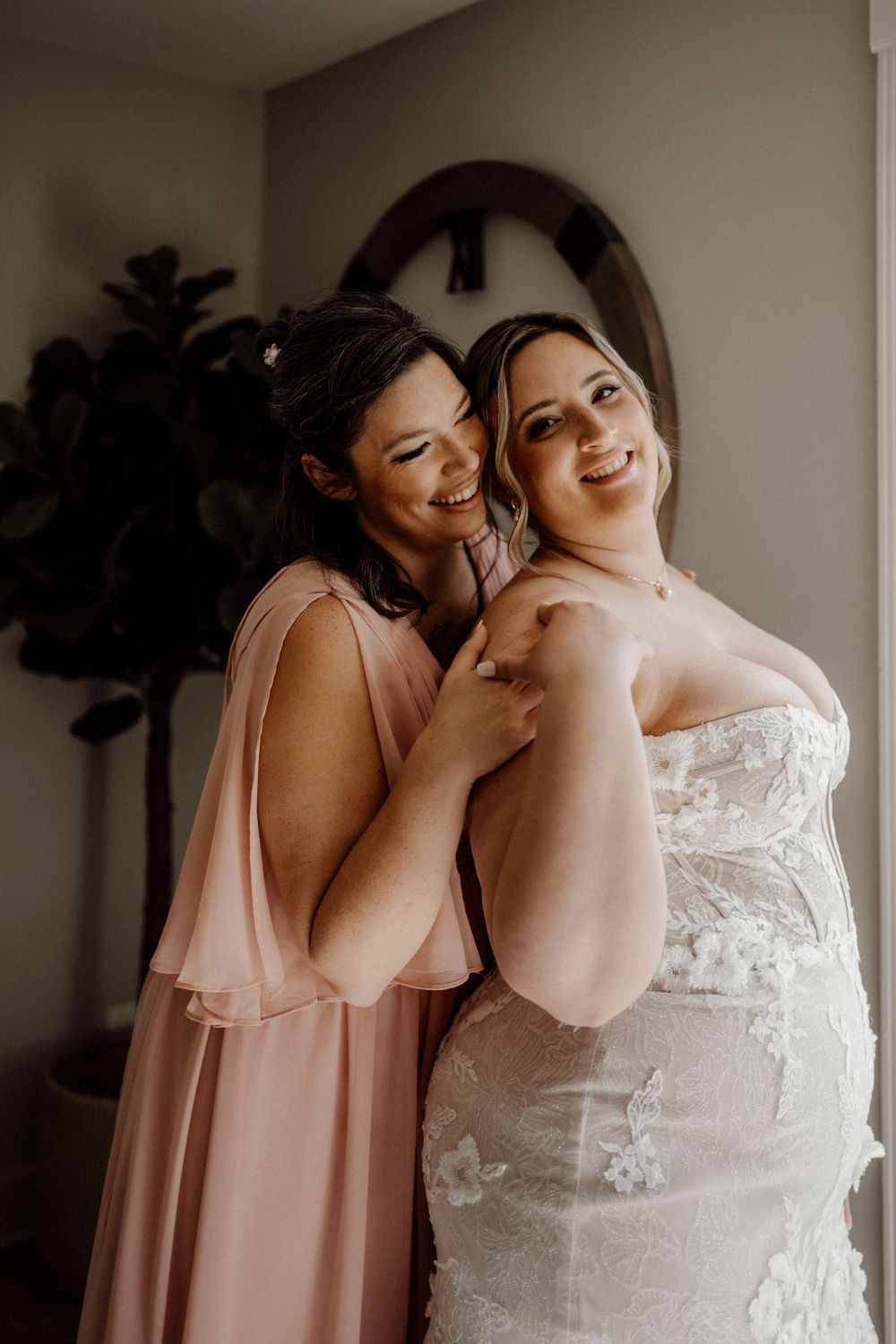 Intimate getting ready moment showing bridesmaids in pink dresses with bride in lace gown.