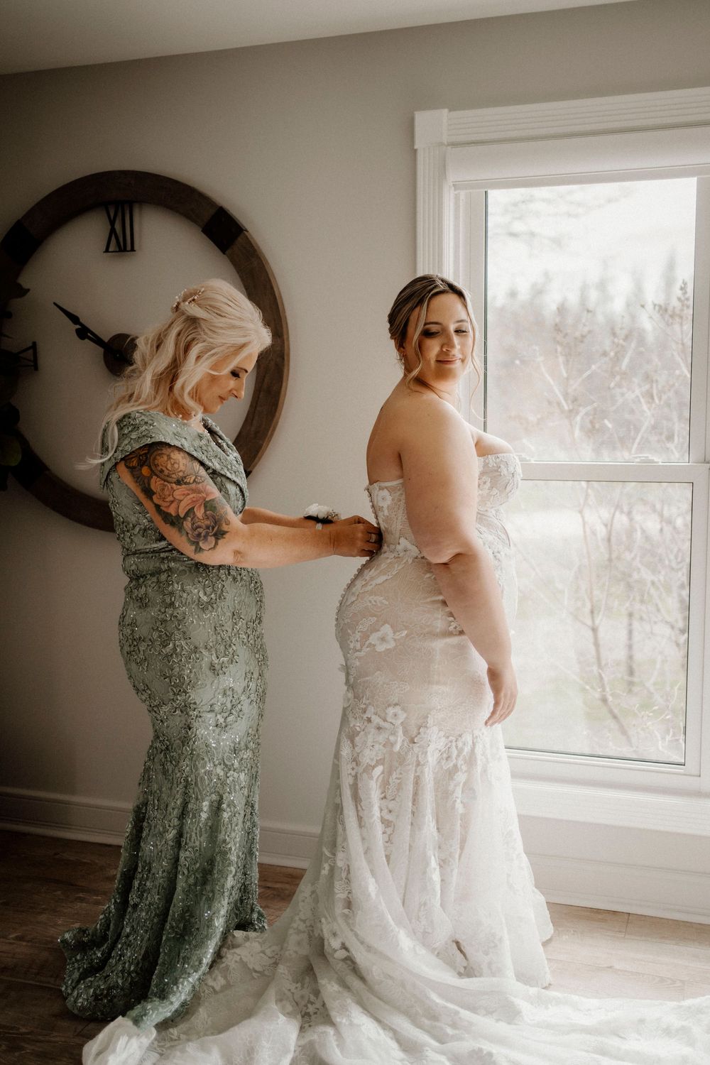 Someone helps with the final adjustments and buttons on a beautiful white lace wedding dress in a sunlit room.