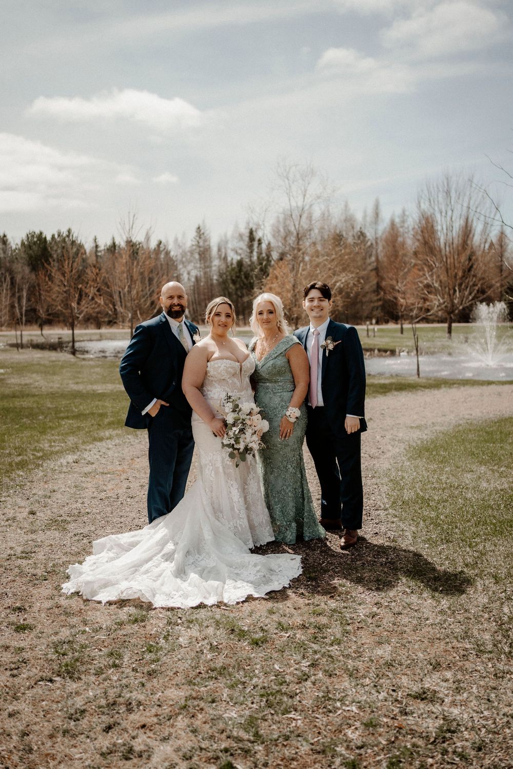 Small wedding group poses for photos outdoors on grassy field with trees in background.