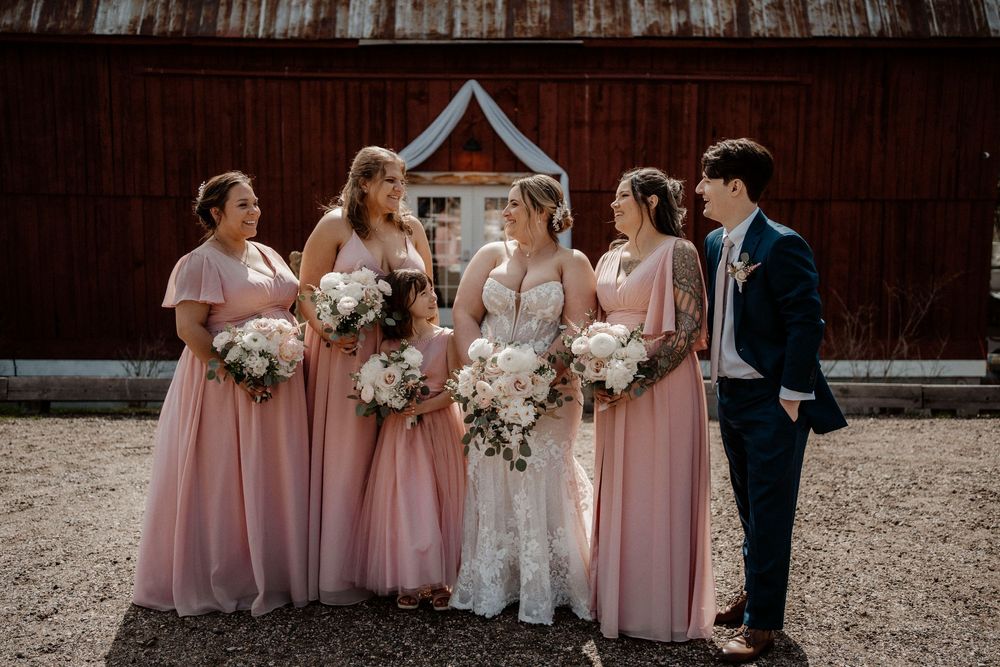 Bridal party in blush pink dresses gathers outside rustic barn venue holding white floral bouquets.