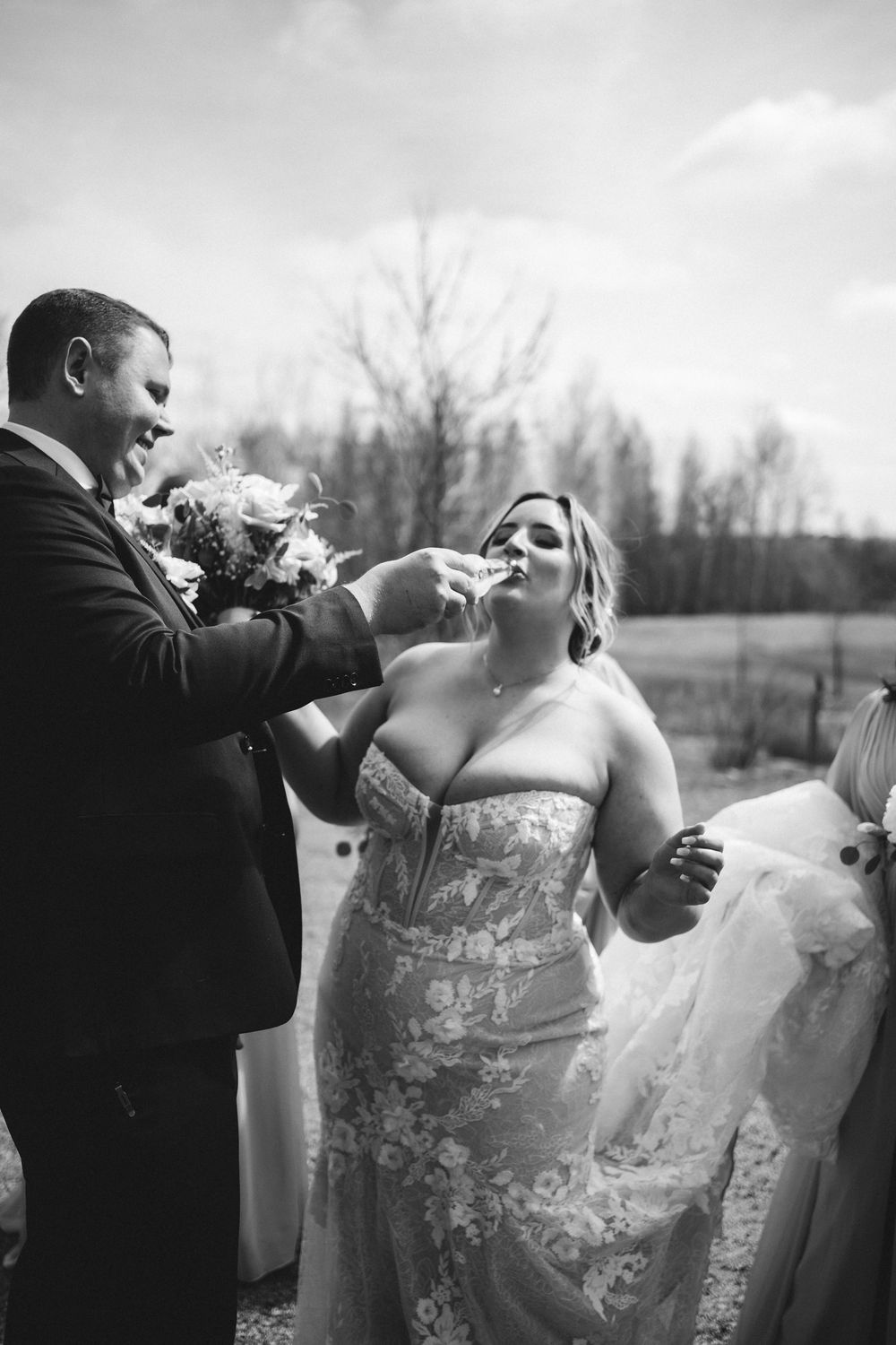 Couple shares playful champagne toast moment during outdoor wedding celebration.