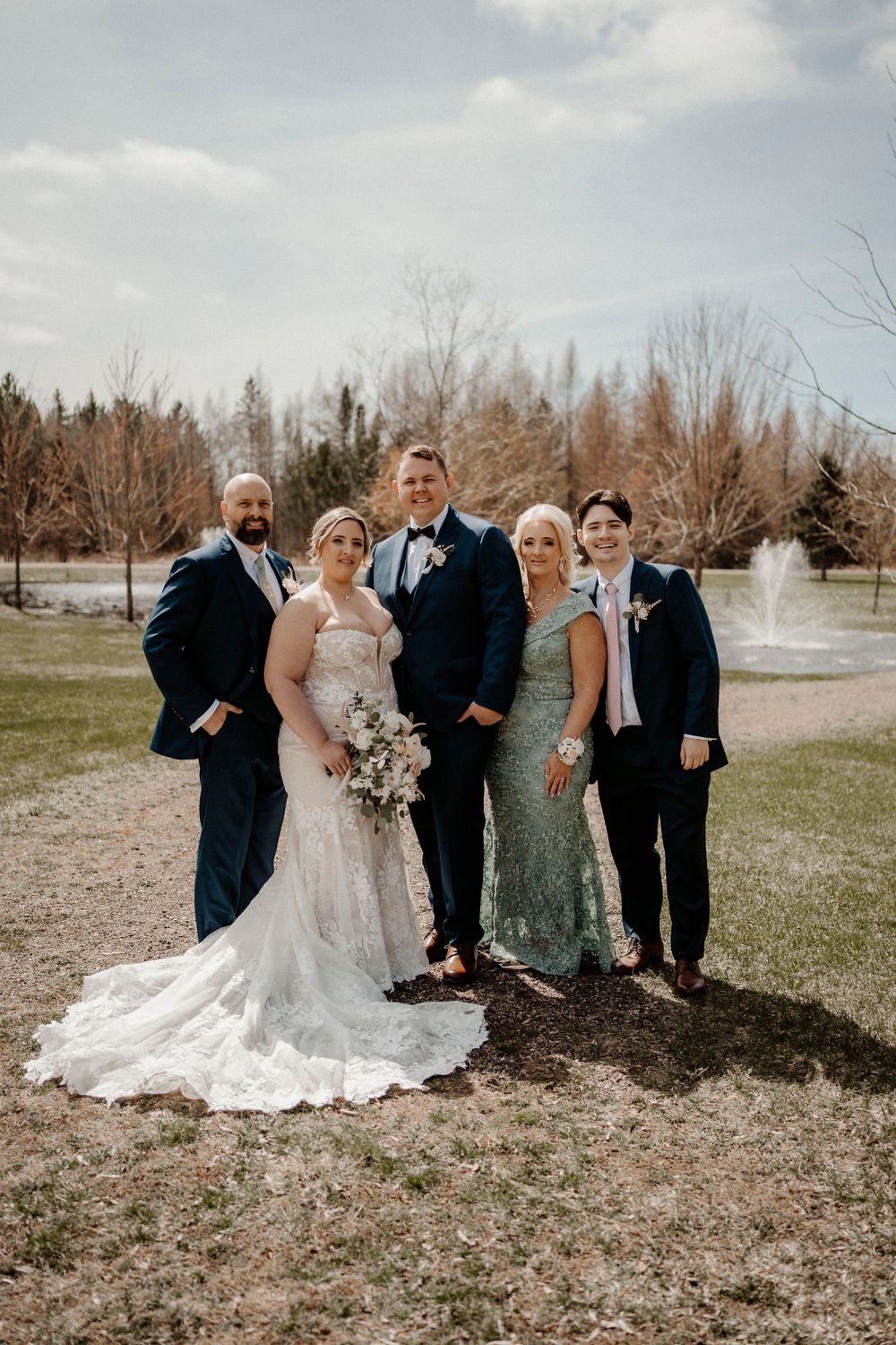 Wedding party stands in a row outdoors on grass with evergreen trees in the background on a cloudy day.