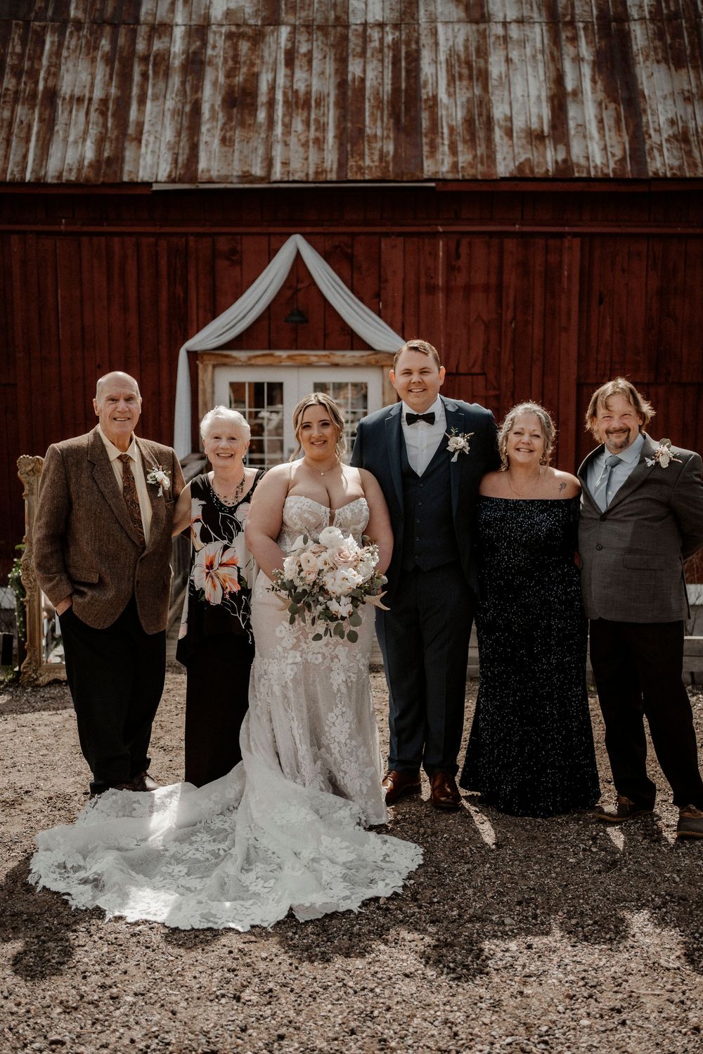 Wedding party poses in front of rustic red barn on overcast day.