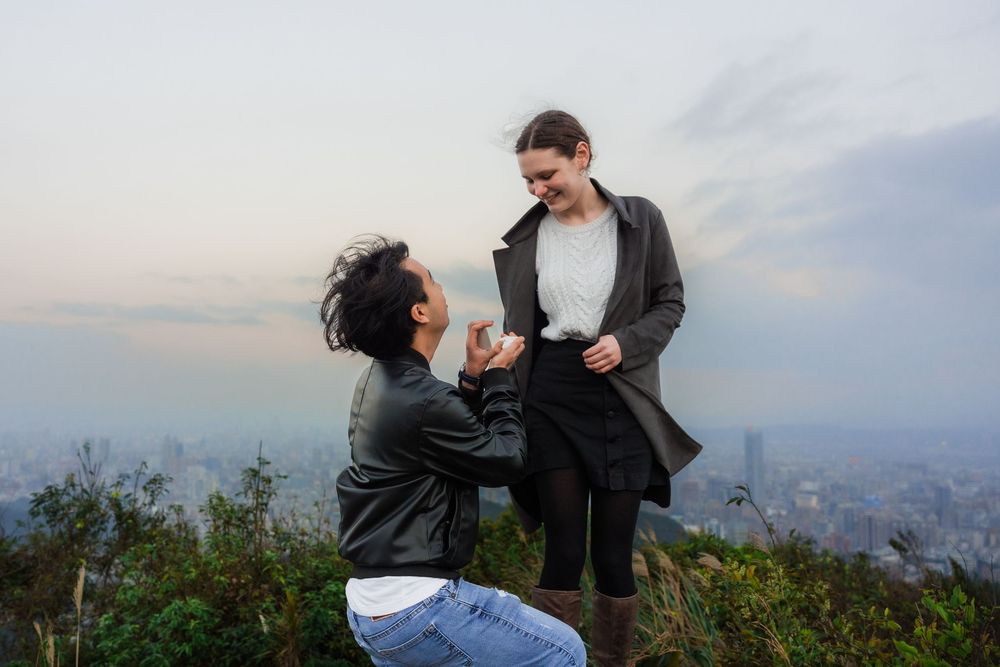 A man kneels on the ground proposing to his girlfriend from a Thumb Lookout Mountain at sunset.