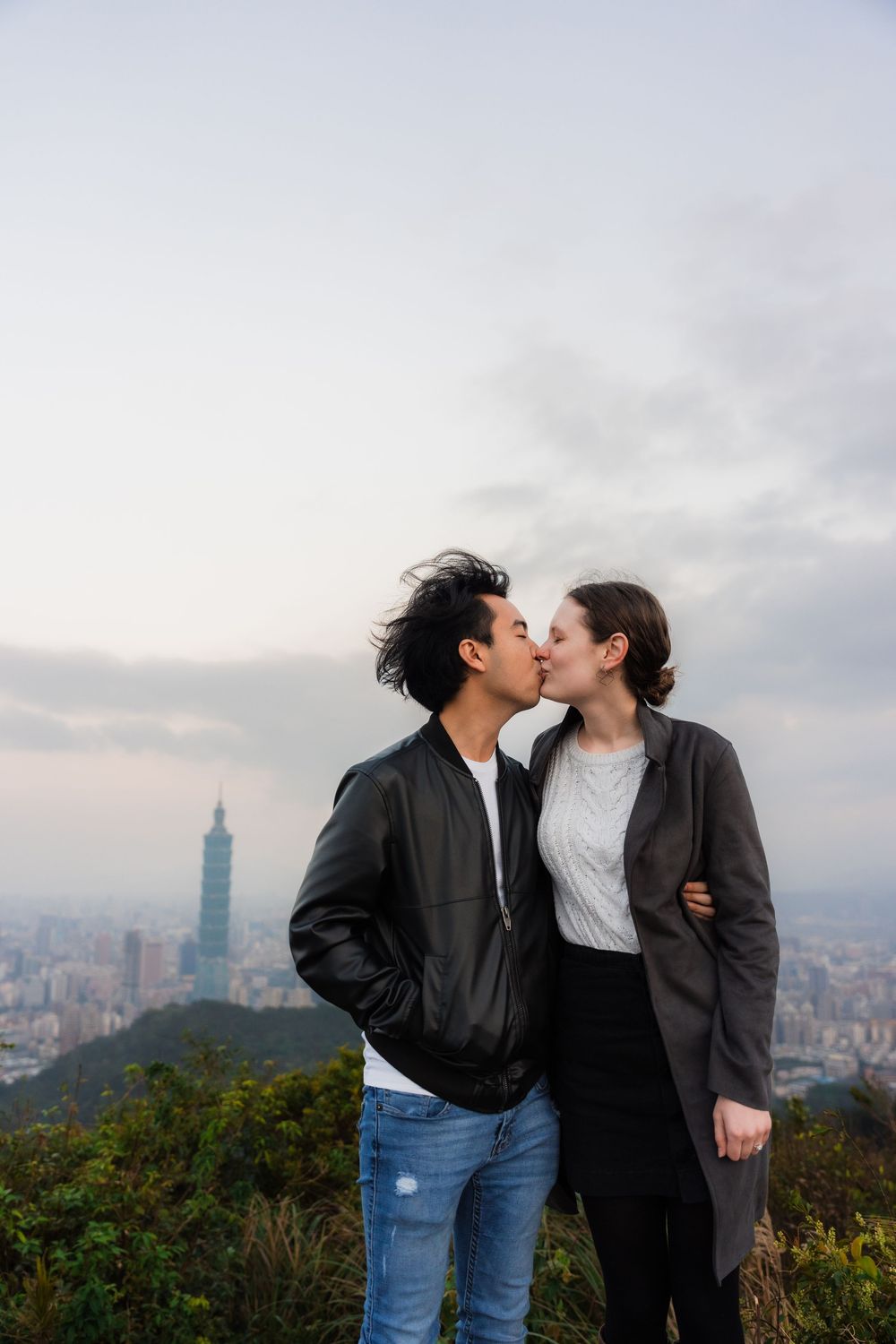 Newly engaged couple sharing a kiss on top Thumb Lookout Mountain with the Taipei 101 skyline in the background.
