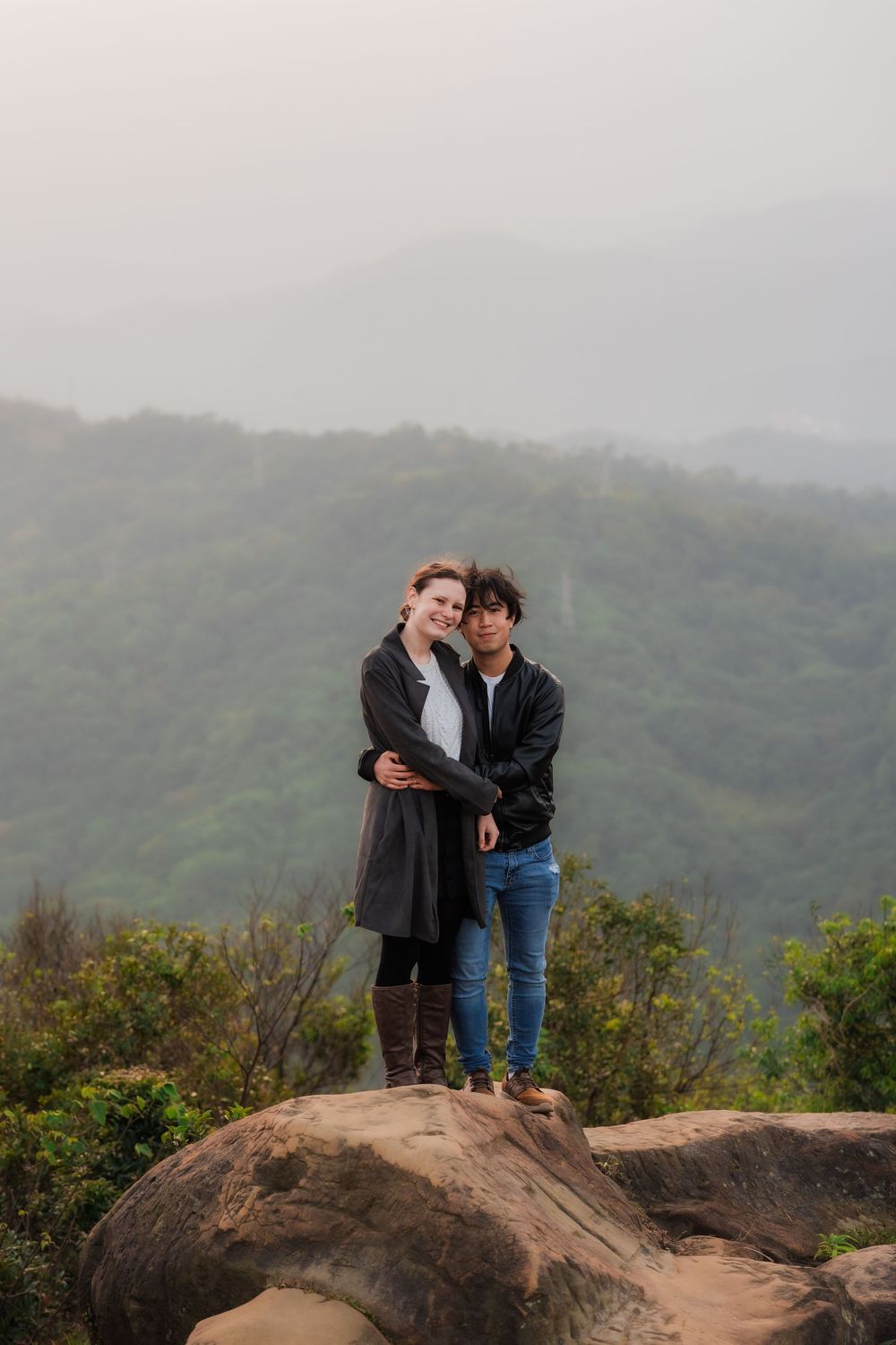 A couple shares an embrace while standing on large rocks with misty mountain scenery in the background.