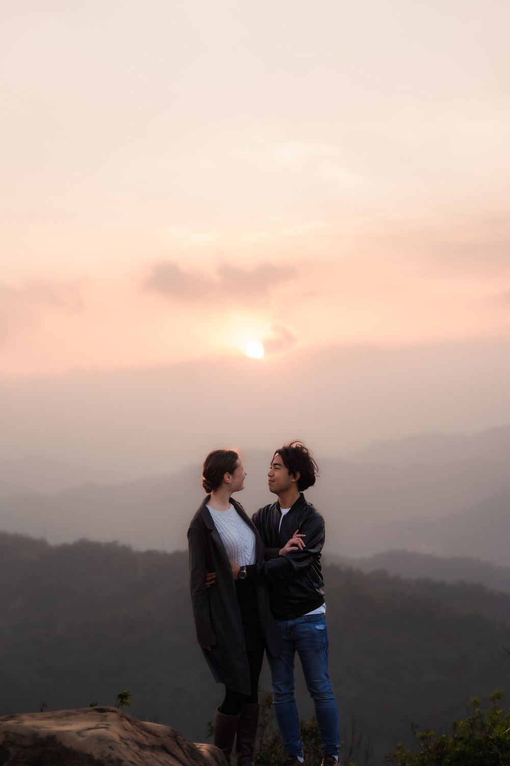 A man and woman in the distance embrace while standing on a high mountain outlook during sunset.