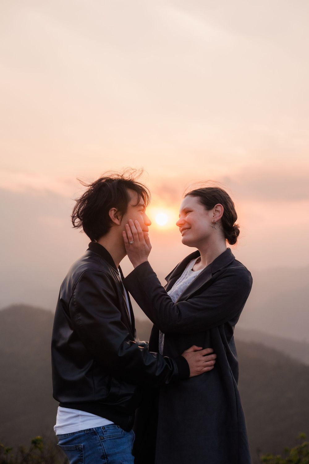 A woman lovingly embraces her partners face while the sun sets over a misty Taiwan mountain setting.
