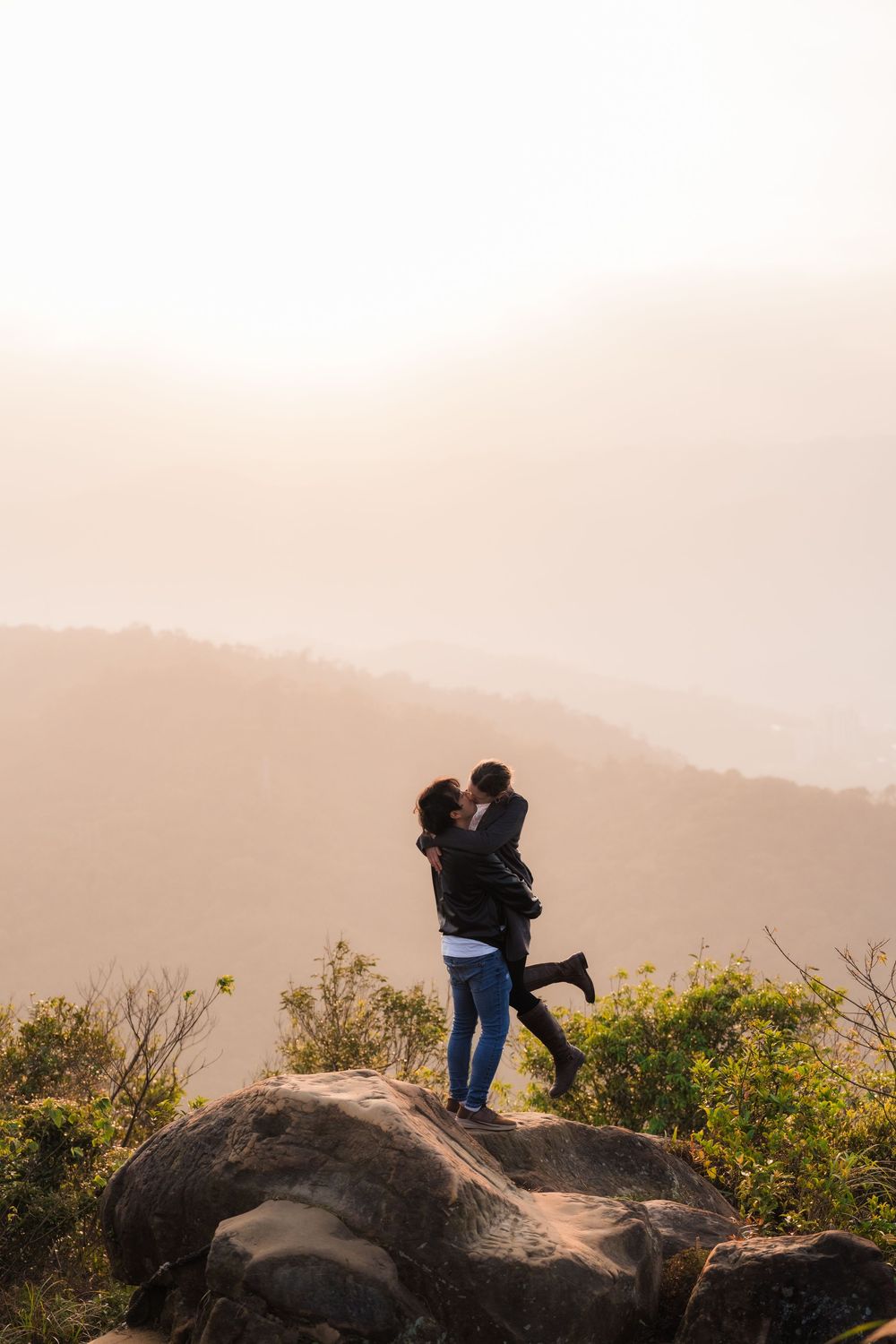 A couple in silhouette  embrace on a hilltop boulder with Taiwan mountains and warm sunset in the background.