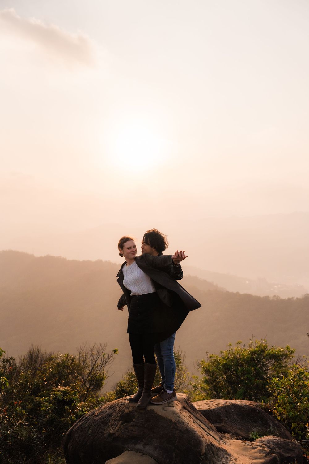 A women is embraced by her partner from behind while the sun sets beyond the misty Taiwan hills in the distance at Thumb Lookout Mountain.
