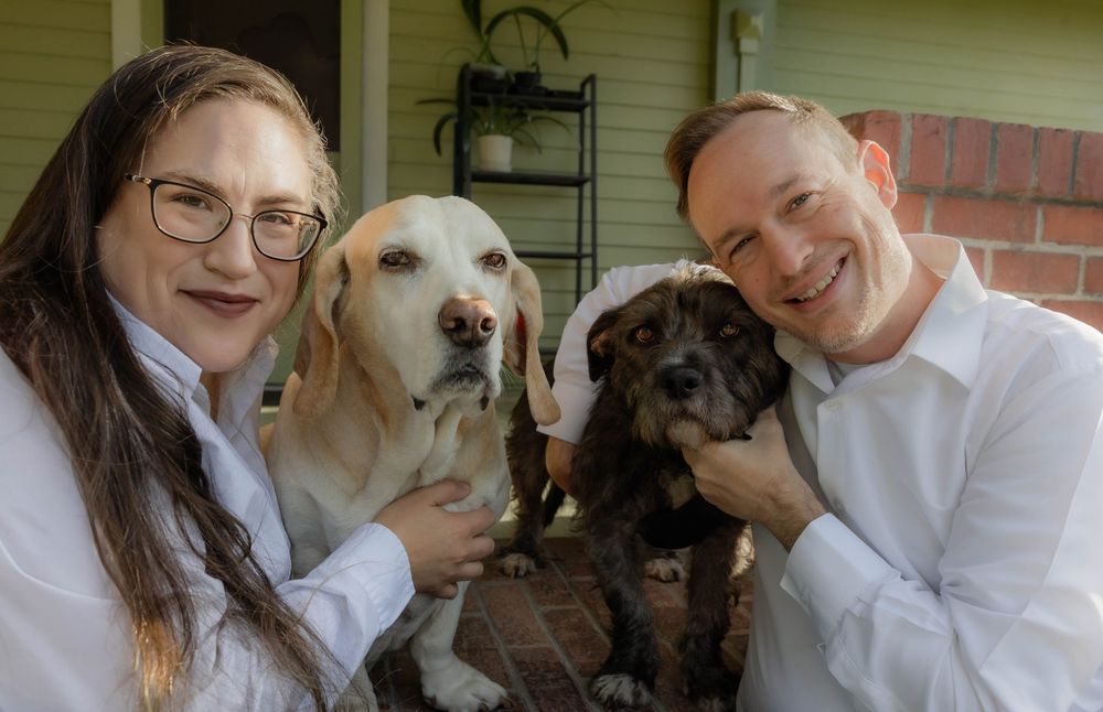 People and dogs pose together for a casual indoor photo near a white-sided house.