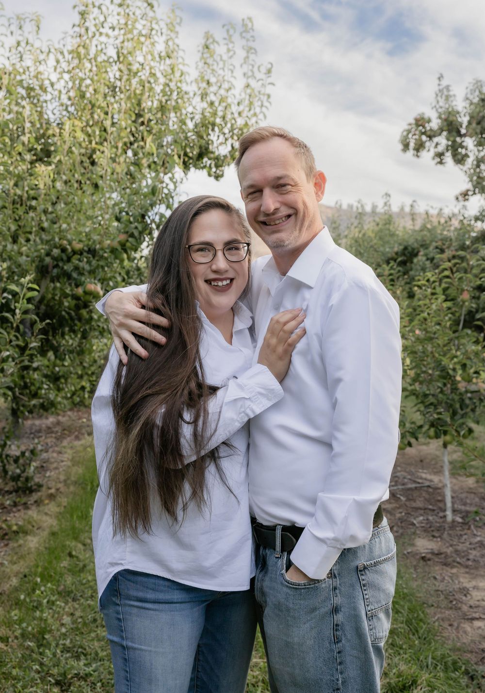 Candid outdoor photoshoot showing joyful moments between two people in white shirts against an orchard backdrop.