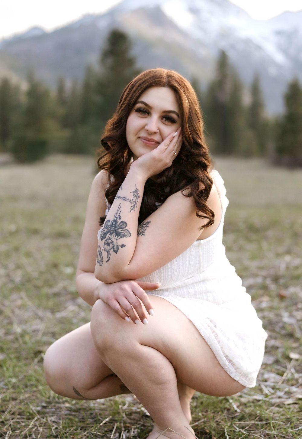 A portrait in a white dress sitting in a mountain meadow with visible tattoos and long curled hair.