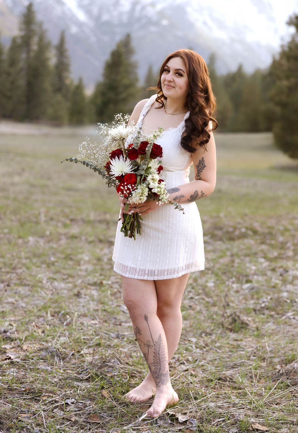 Standing in mountain field holding red and white bouquet wearing short white dress.